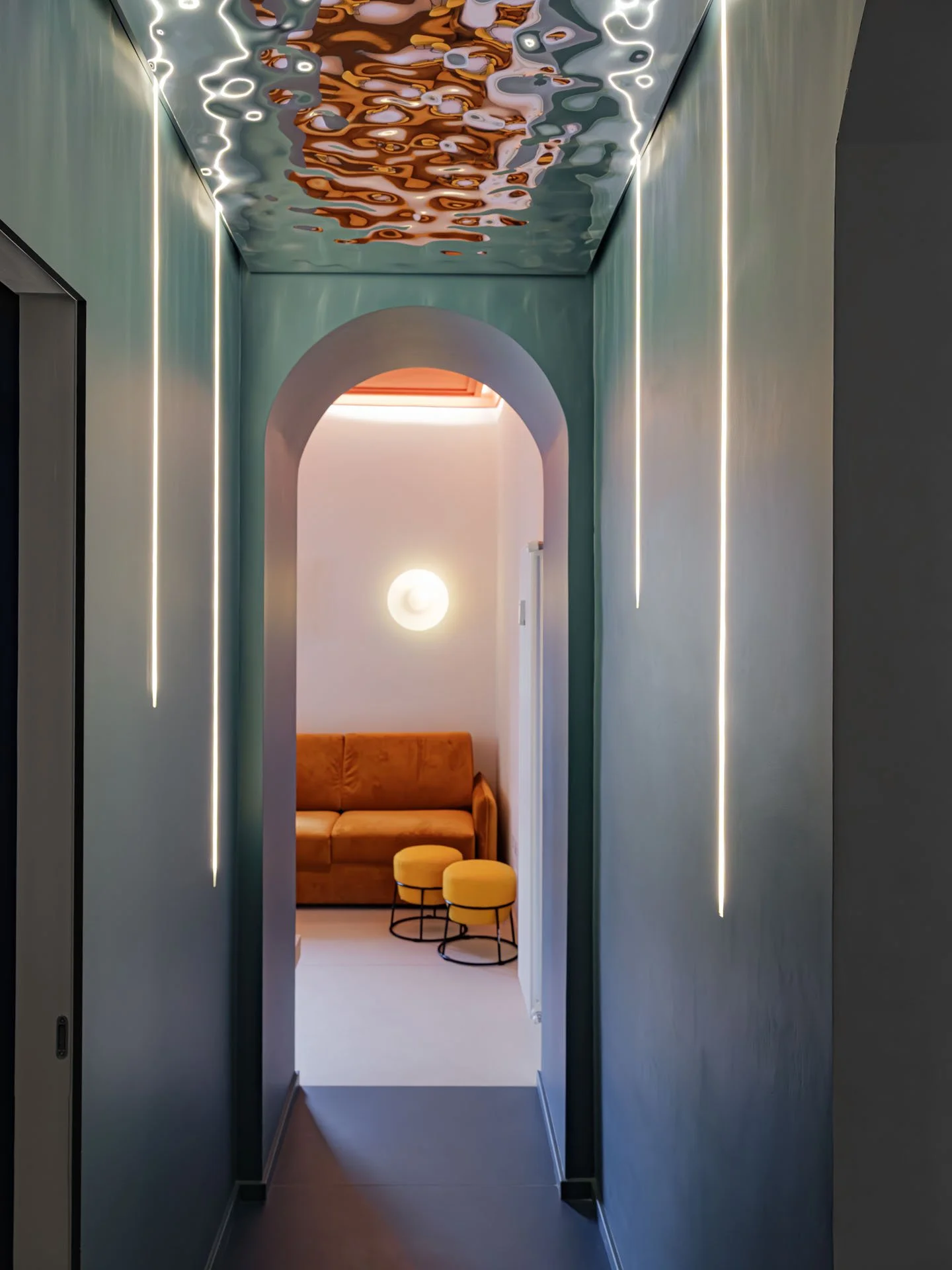 Un corridoio moderno con pareti verdi e illuminazione a LED, conduce a un'area salotto con un divano arancione e due sgabelli gialli.