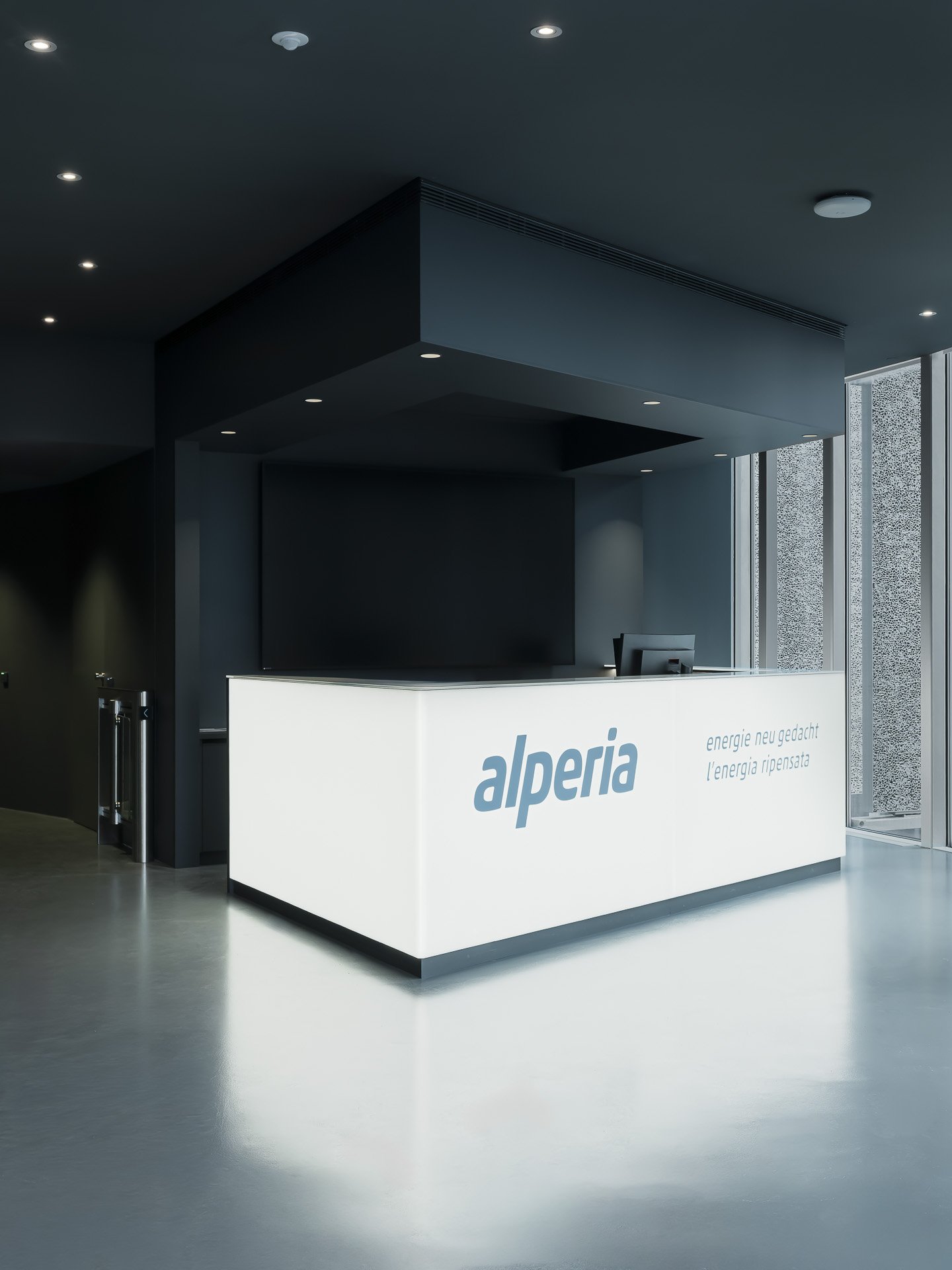 Reception area with a white counter displaying the word "alperia" in blue, and Italian slogans 'energie nel gadgets' and 'l'energia ripensata', in a modern building with dark walls and large windows.