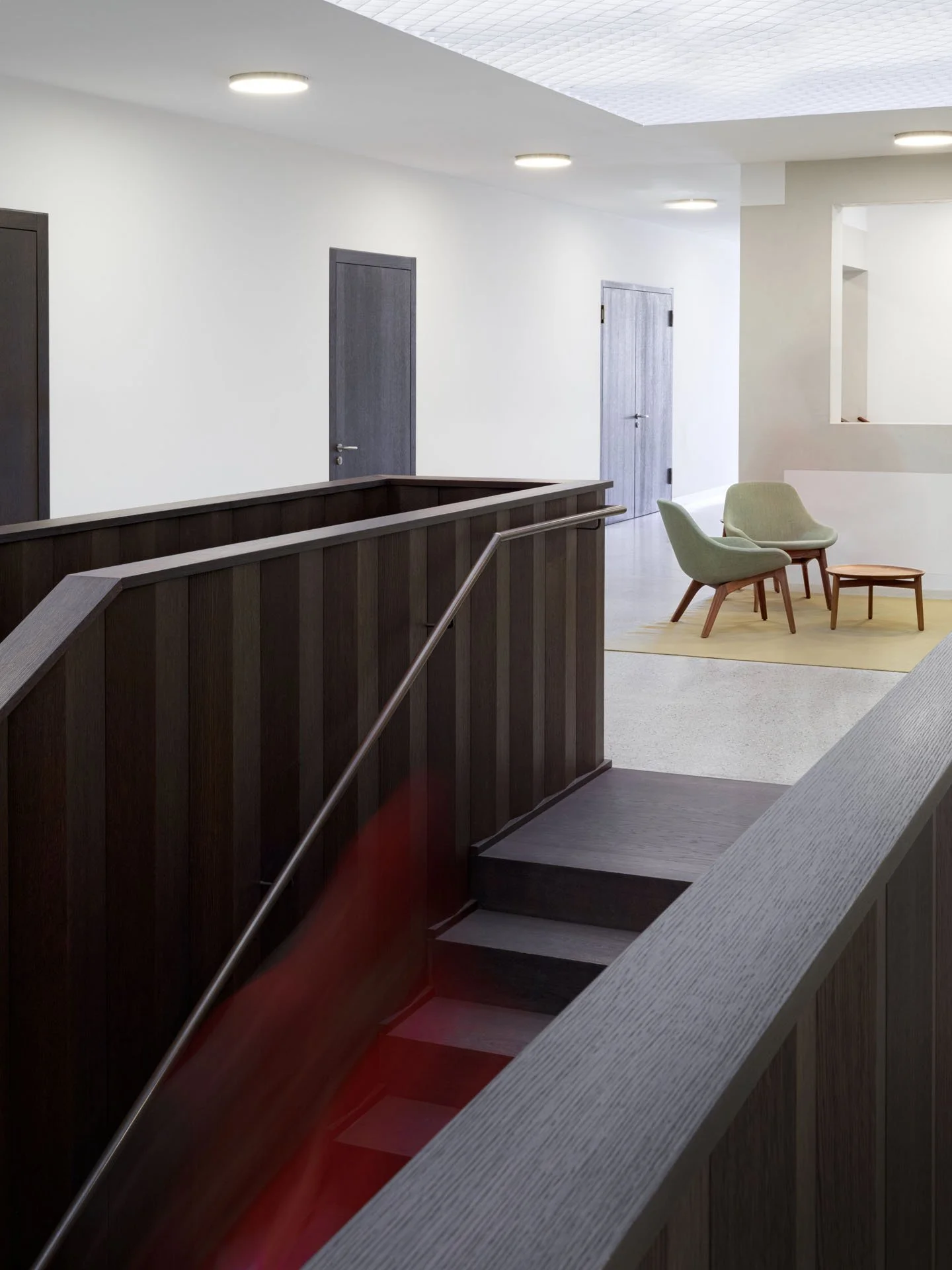 Ingresso di un edificio con sedie bianche e tavolino, pareti bianche, porte di legno grigio e soffitto con luci rotonde.