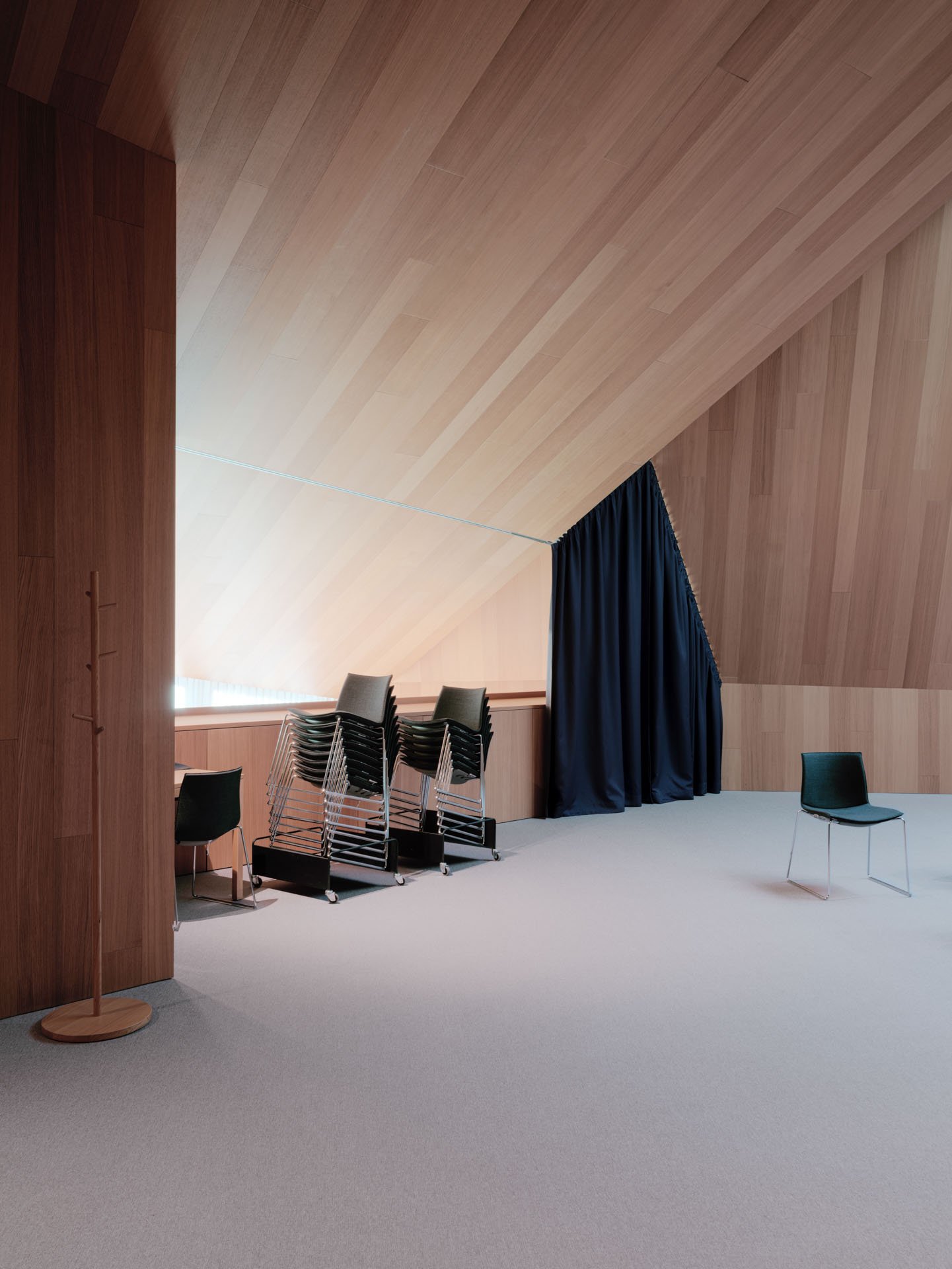 Interno di una sala con pareti e soffitto in legno, alcune sedie impilate e una sedia singola, un tendone nero e un appendiabiti vuoto.