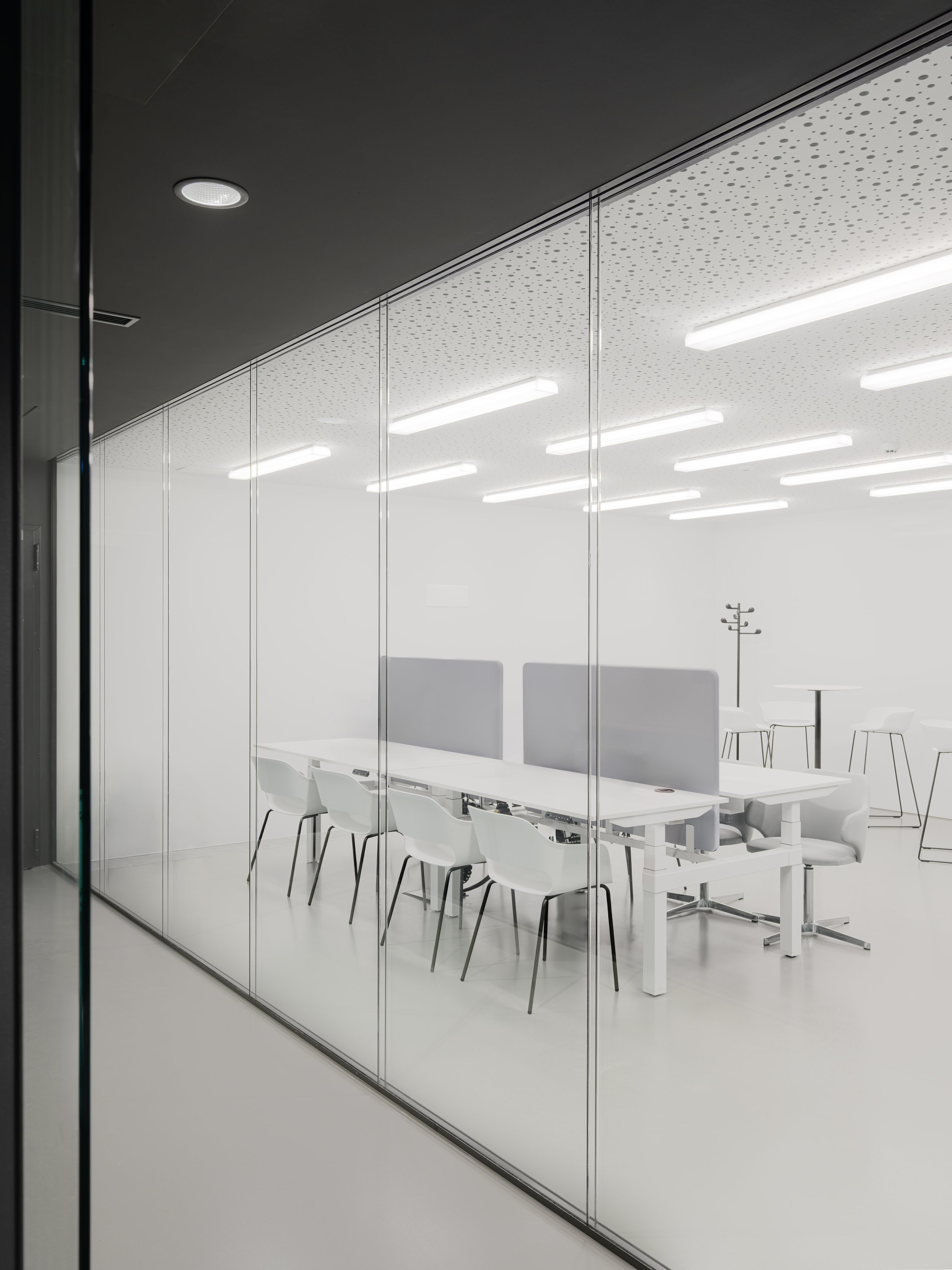 Vista di un ufficio moderno visto attraverso una parete di vetro, con sedie bianche e tavoli, illuminazione a soffitto con luci a LED, pareti biene e arredi minimalisti.