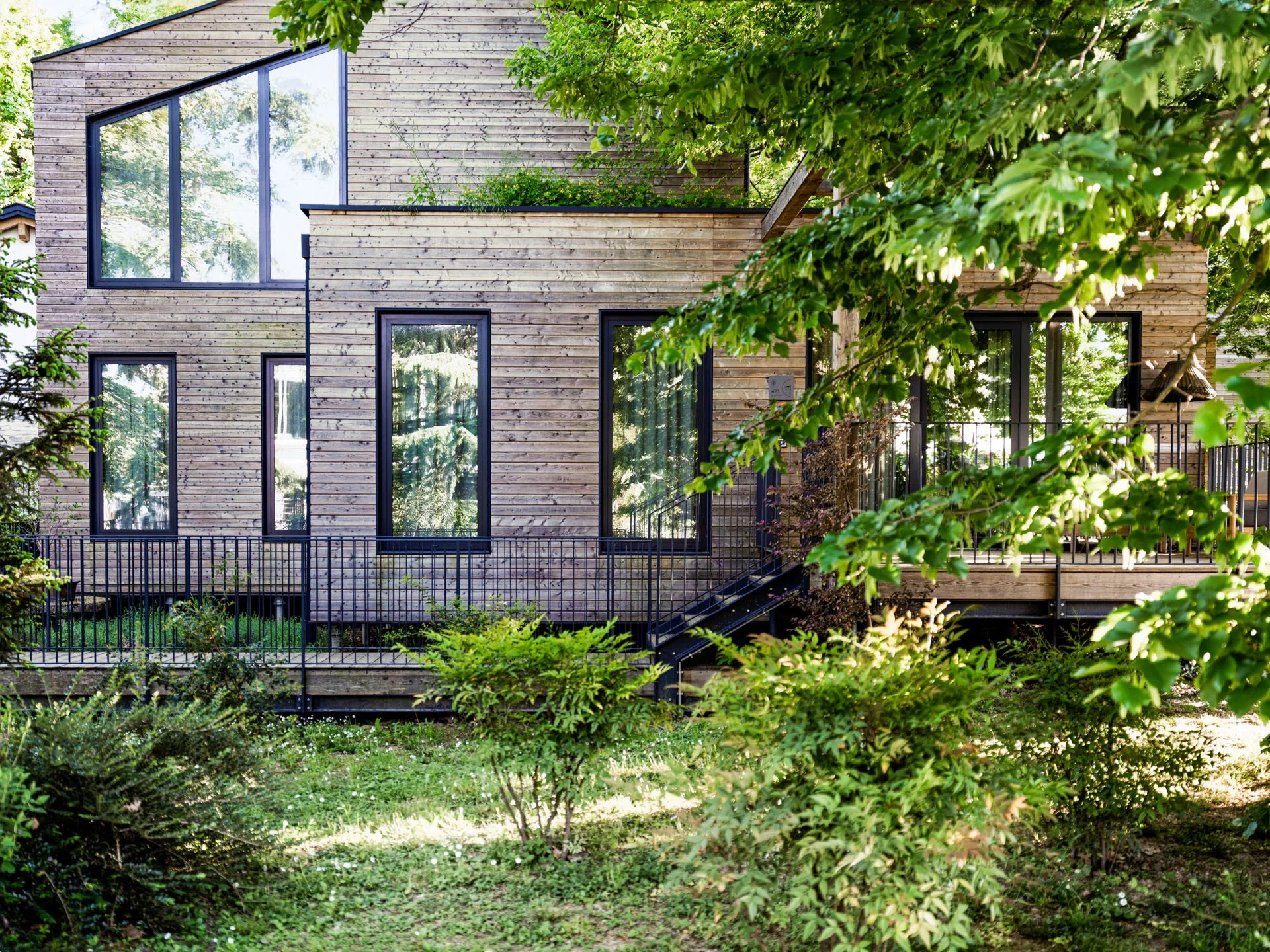 Casa moderna in legno con grandi finestrini e balcone, circondata da alberi e vegetazione verde.