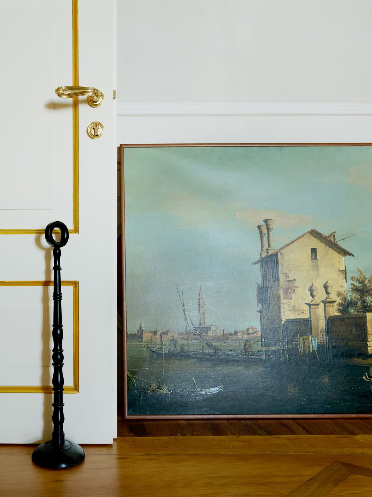 Interno di una stanza con un quadro che rappresenta un paesaggio marittimo e una porta bianca con maniglia e serratura in ottone, accompagnati da un lampione nero.