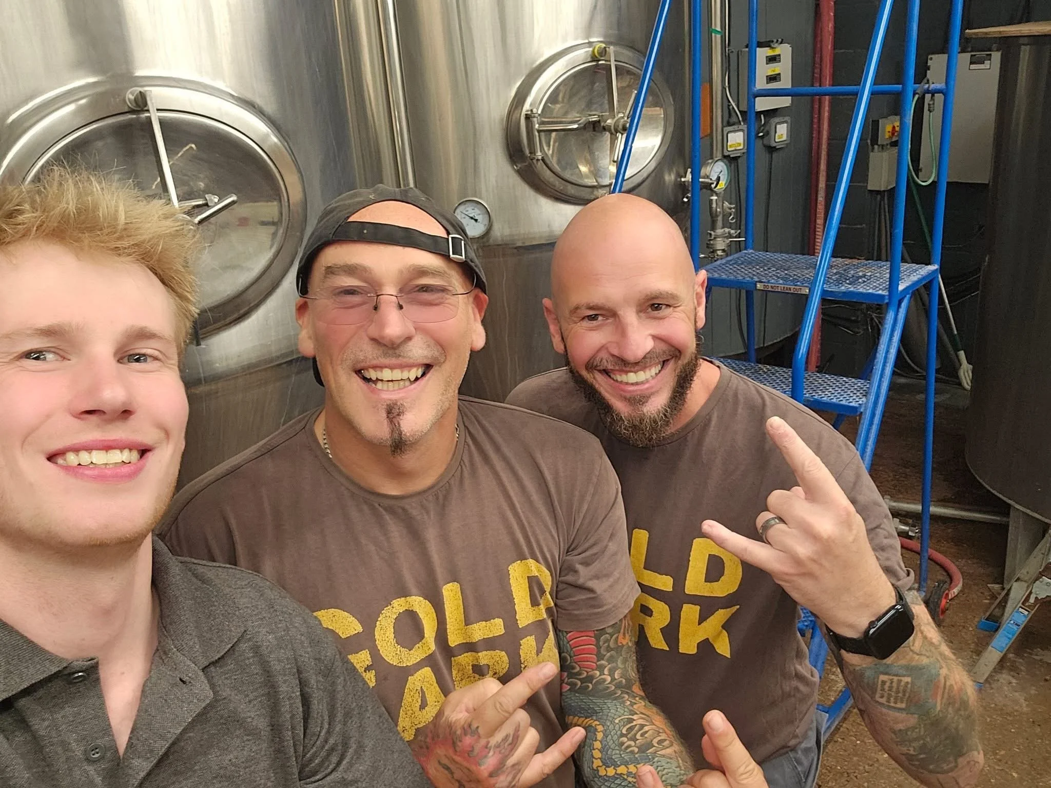 Three smiling men taking a selfie in front of large stainless steel tanks in a brewery or industrial setting.