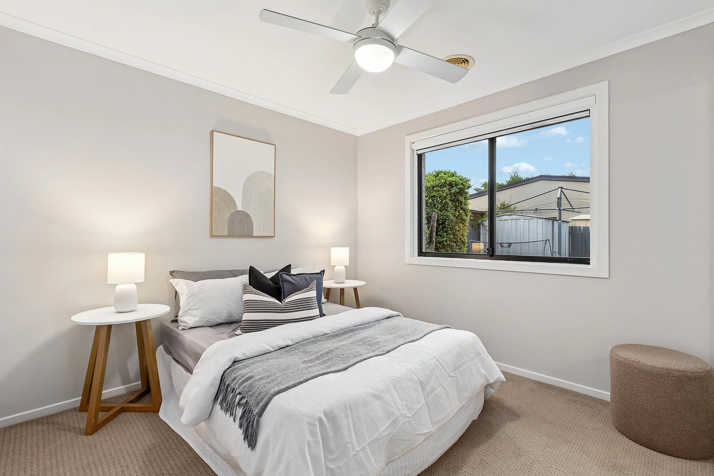 Bedroom with a bed, two bedside tables with lamps, a window showing an outdoor scene with trees and neighboring buildings, light-colored walls, ceiling fan, beige carpet, wall art above the bed, and a round ottoman.