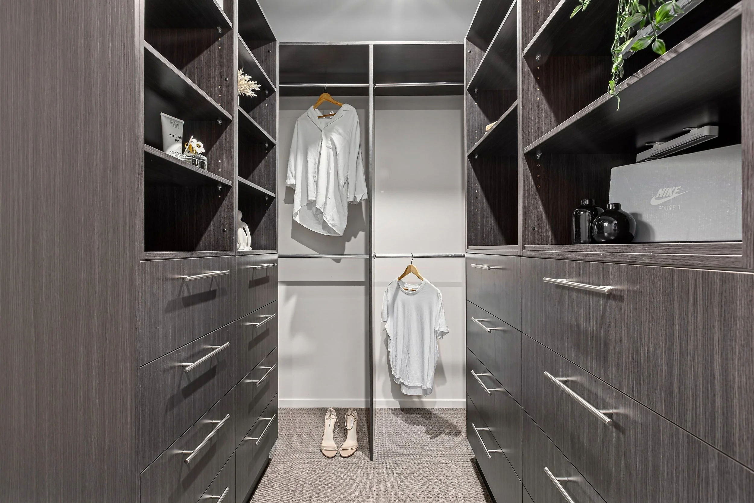 A walk-in closet with dark wood shelves and drawers, white hanging shirts, beige high heels, and a gray Nike box.
