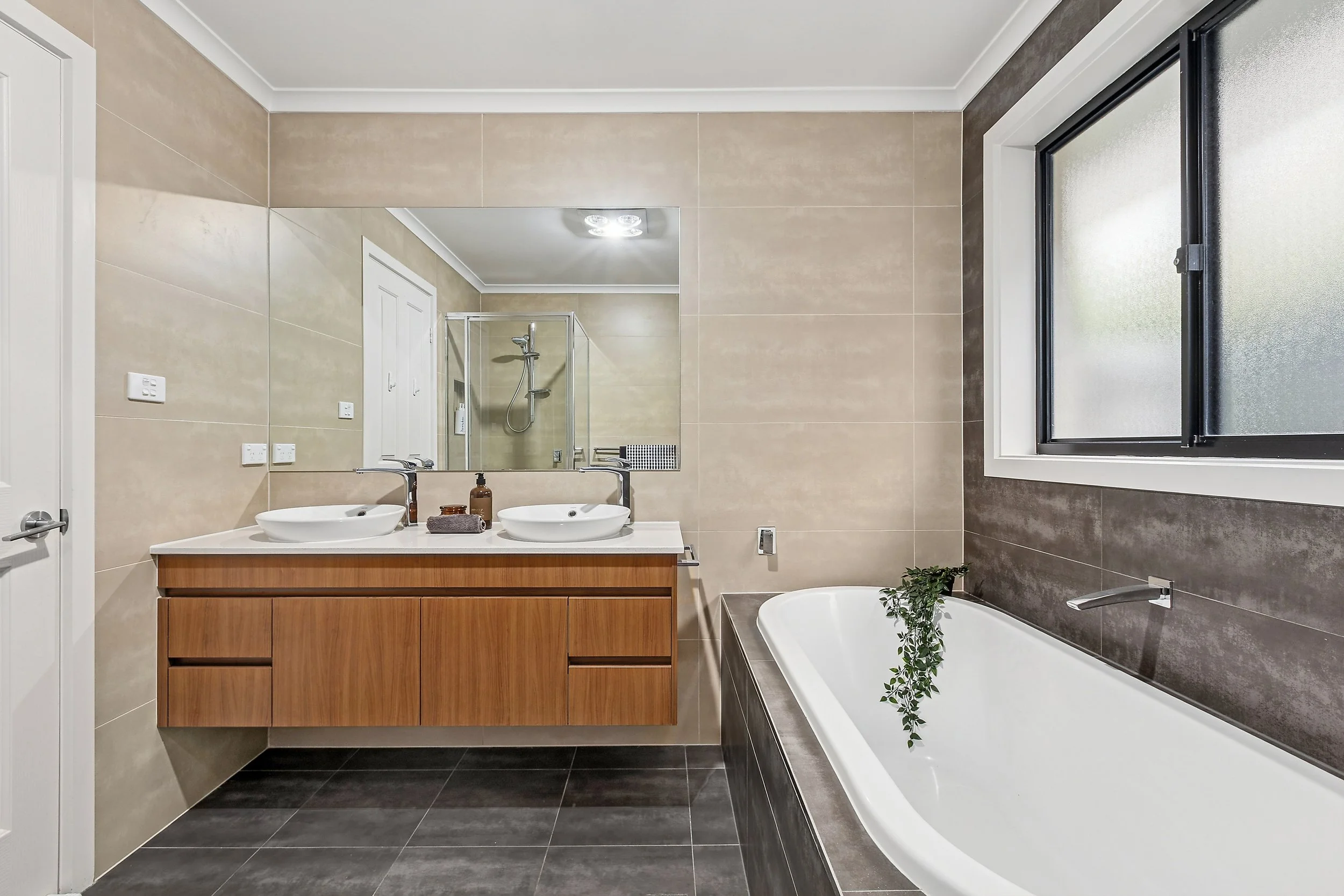 Modern bathroom with beige tiled walls, a large mirror, dual white vessel sinks on a wooden vanity, a bathtub with a plant inside, and a window with frosted glass.