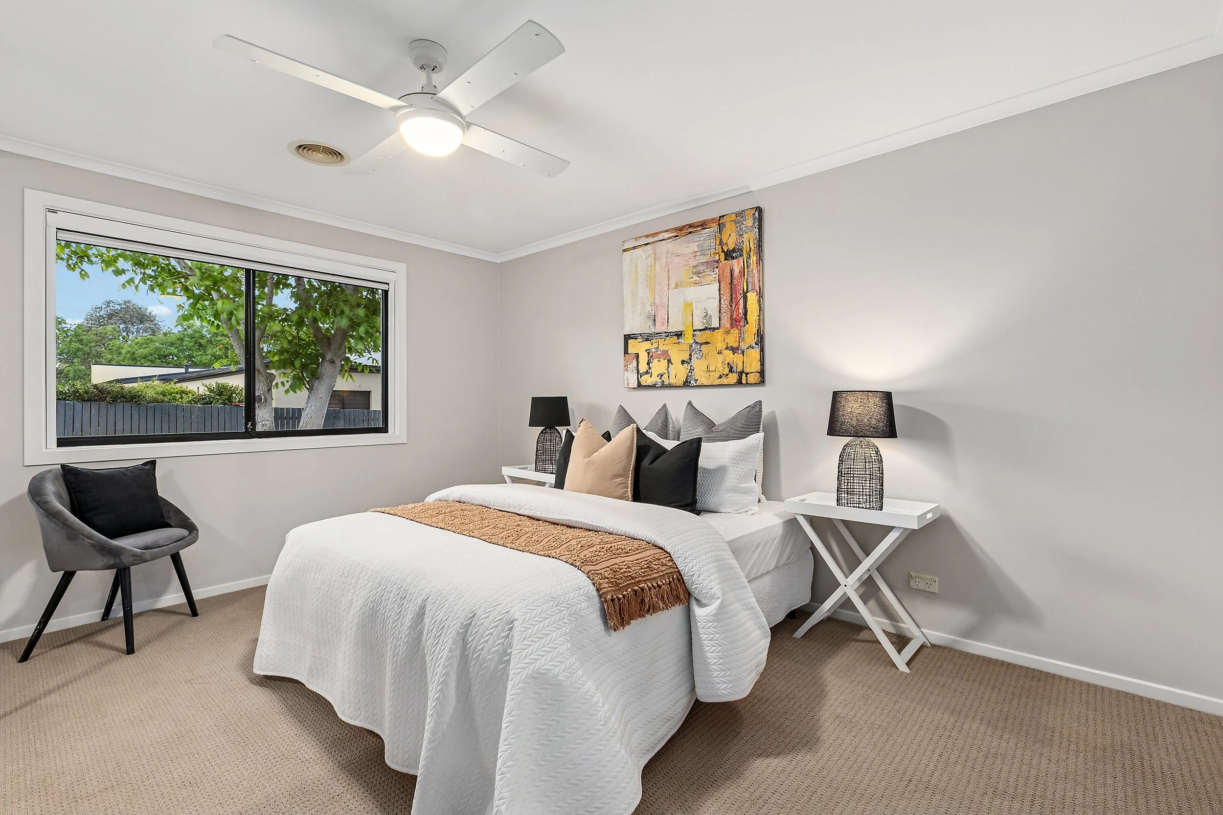 Bedroom with white bedding, decorative pillows, beige throw blanket, black lamps, abstract artwork, window with view of tree and fence, beige carpet, gray chair with black cushion.