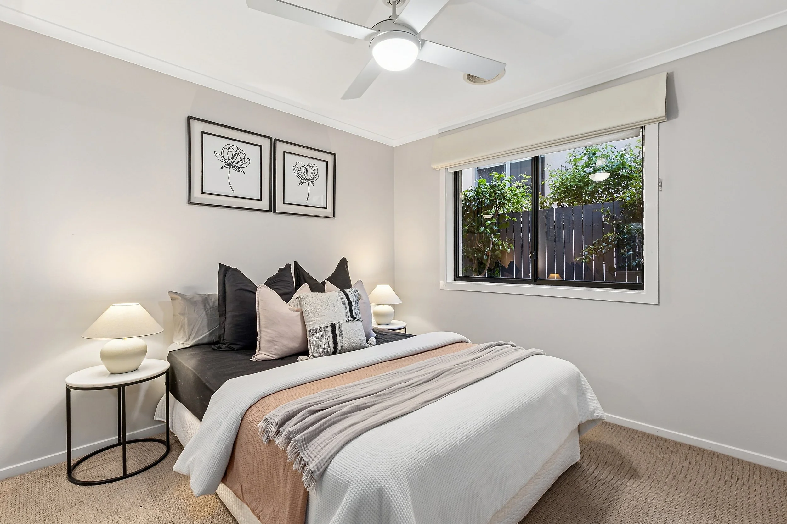 A clean, minimalist bedroom with a bed, nightstands, lamps, and wall art, with a large window showing greenery outside.