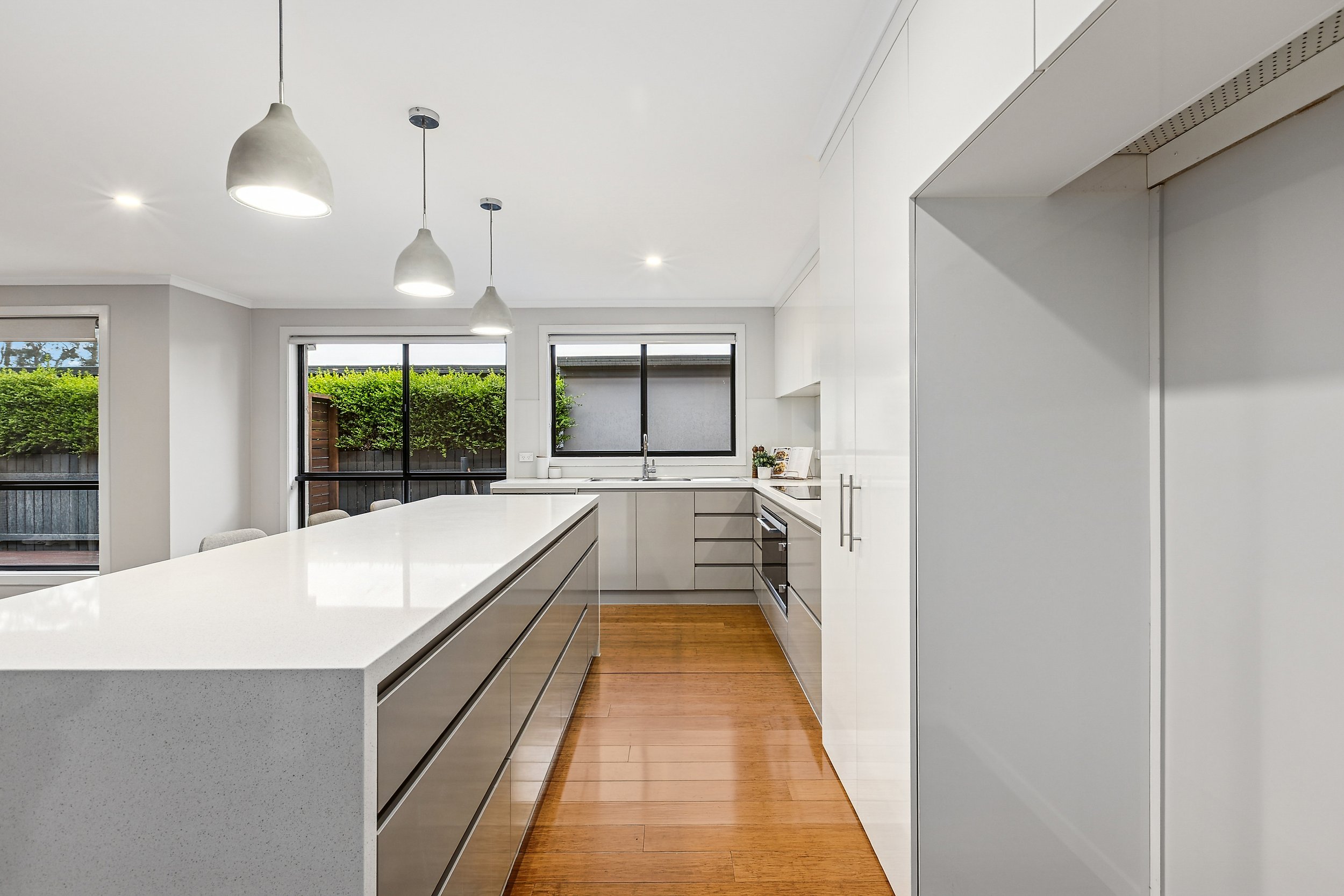 Modern kitchen with white cabinets, a white countertop island, and wooden flooring, illuminated by three hanging pendant lights and large windows showing outdoor greenery.