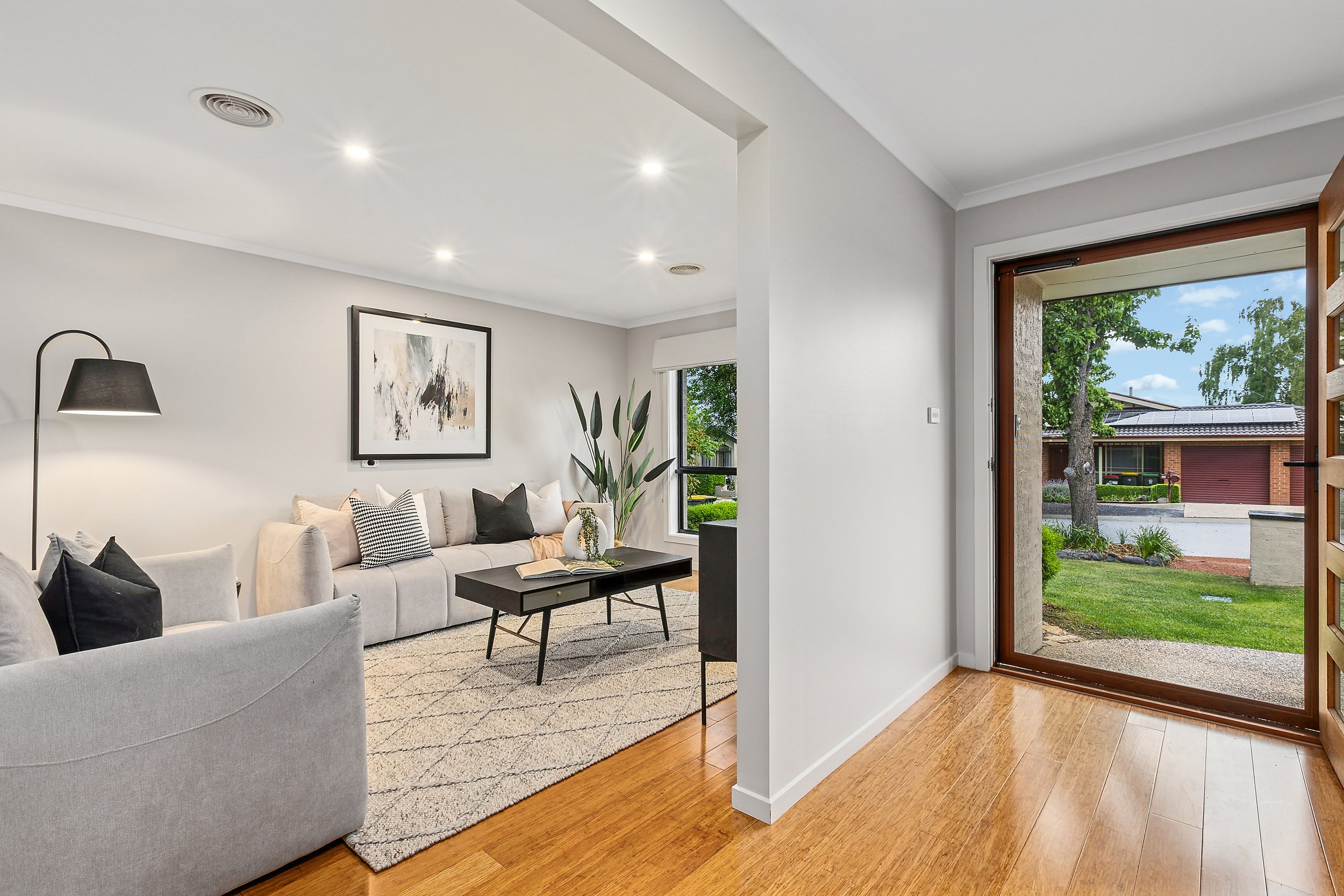 Living room with a beige sofa, black and white decorative pillows, a black coffee table, a side table, a painting on the wall, a floor lamp, and a large potted plant. An open door shows a view of a front yard with green grass, trees, and a neighborin