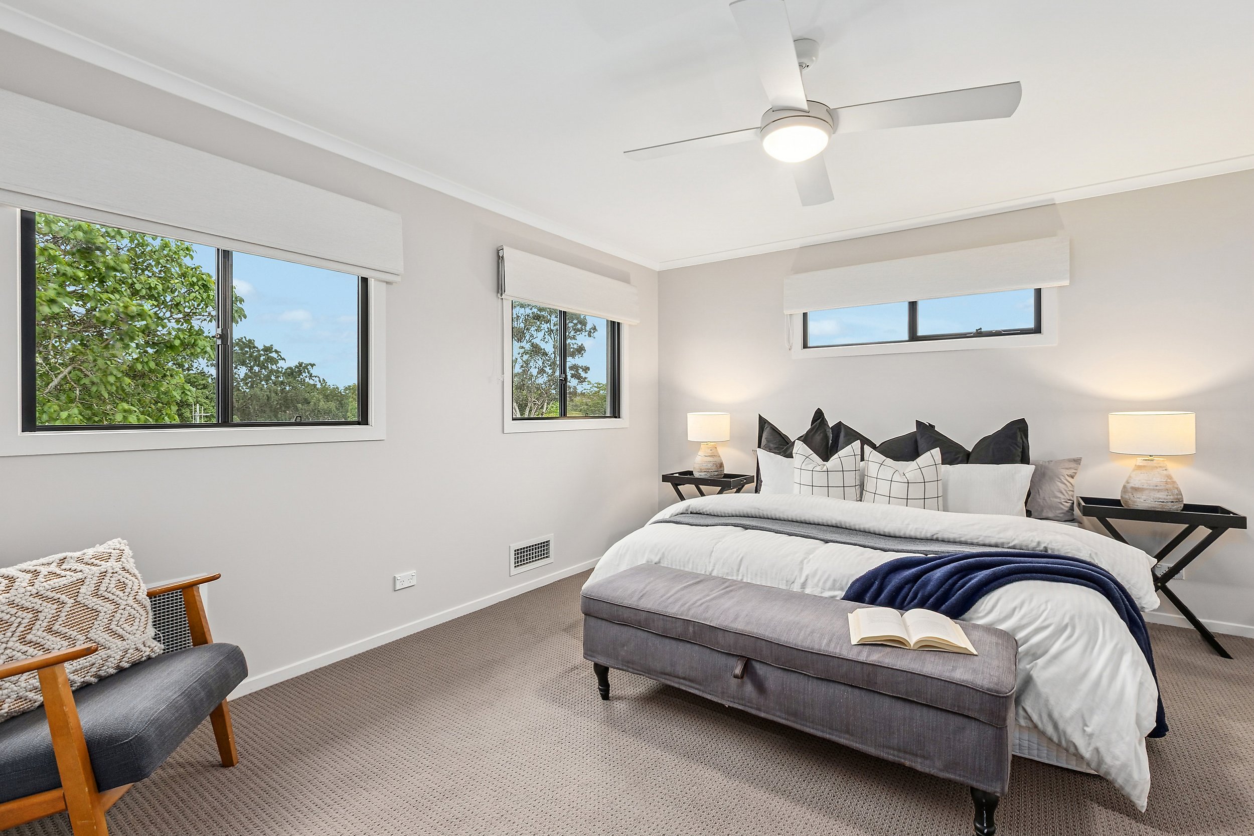 A bedroom with white walls, three windows with white blinds, a bed with black, white, and gray pillows, a gray upholstered bench at the foot of the bed with an open book, two black side tables with lamps, a ceiling fan with lights, and a chair with a
