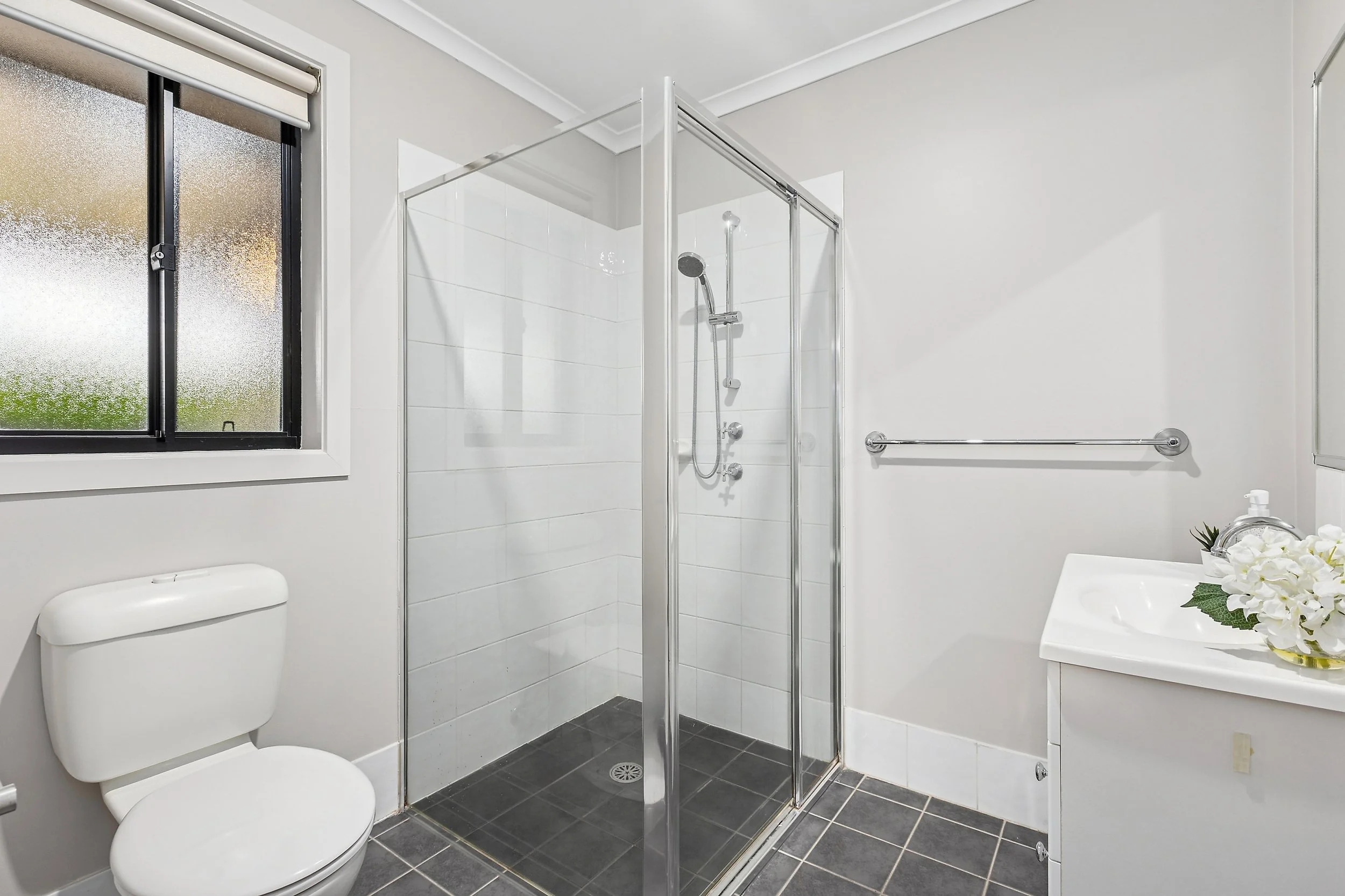 A bathroom with a toilet, a small window, a shower with a glass door, a white sink with a flower arrangement, and a towel bar on the wall.