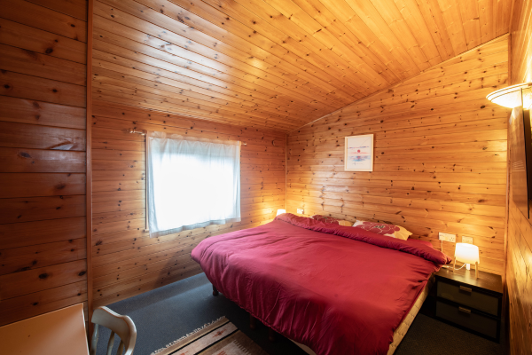 A cozy bedroom with wooden walls and ceiling, a window with white curtains, a large bed with a red bedspread, a nightstand with a lamp, and a framed picture on the wall.