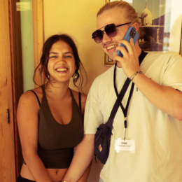 Two smiling young women standing close together, one wearing sunglasses and holding a phone, the other wearing a black tank top, inside a room with yellow walls.