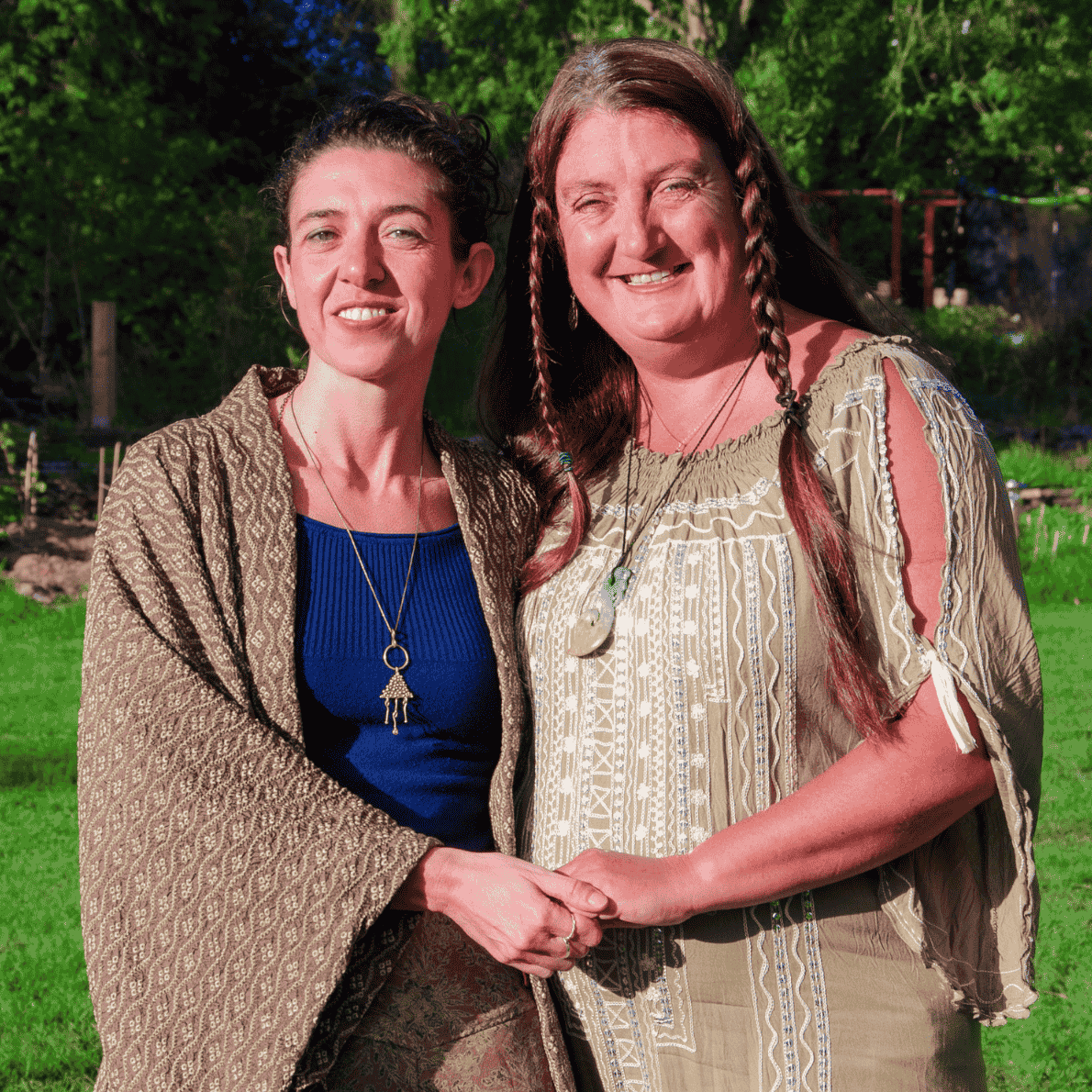 Two women standing outdoors, smiling and holding hands. One woman has dark hair in braids and the other has curly hair. Both are wearing casual, bohemian style clothing with jewelry.