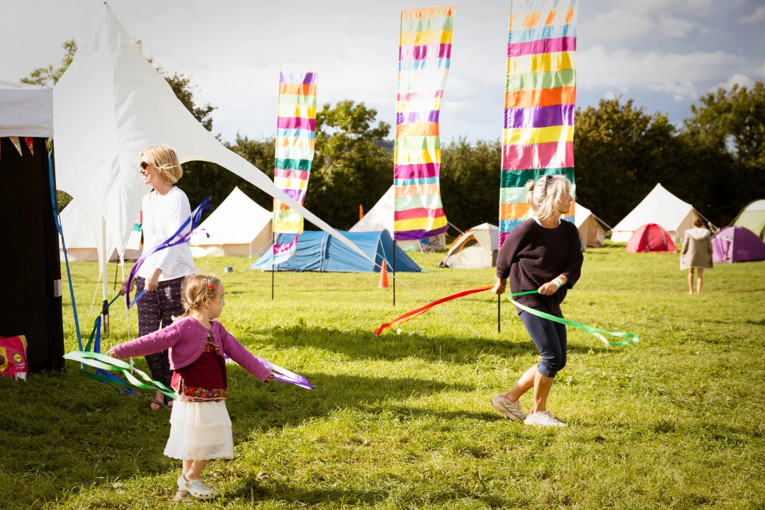 People at a camping site on a grassy field, with colorful banners and tents in the background, enjoying a sunny day.