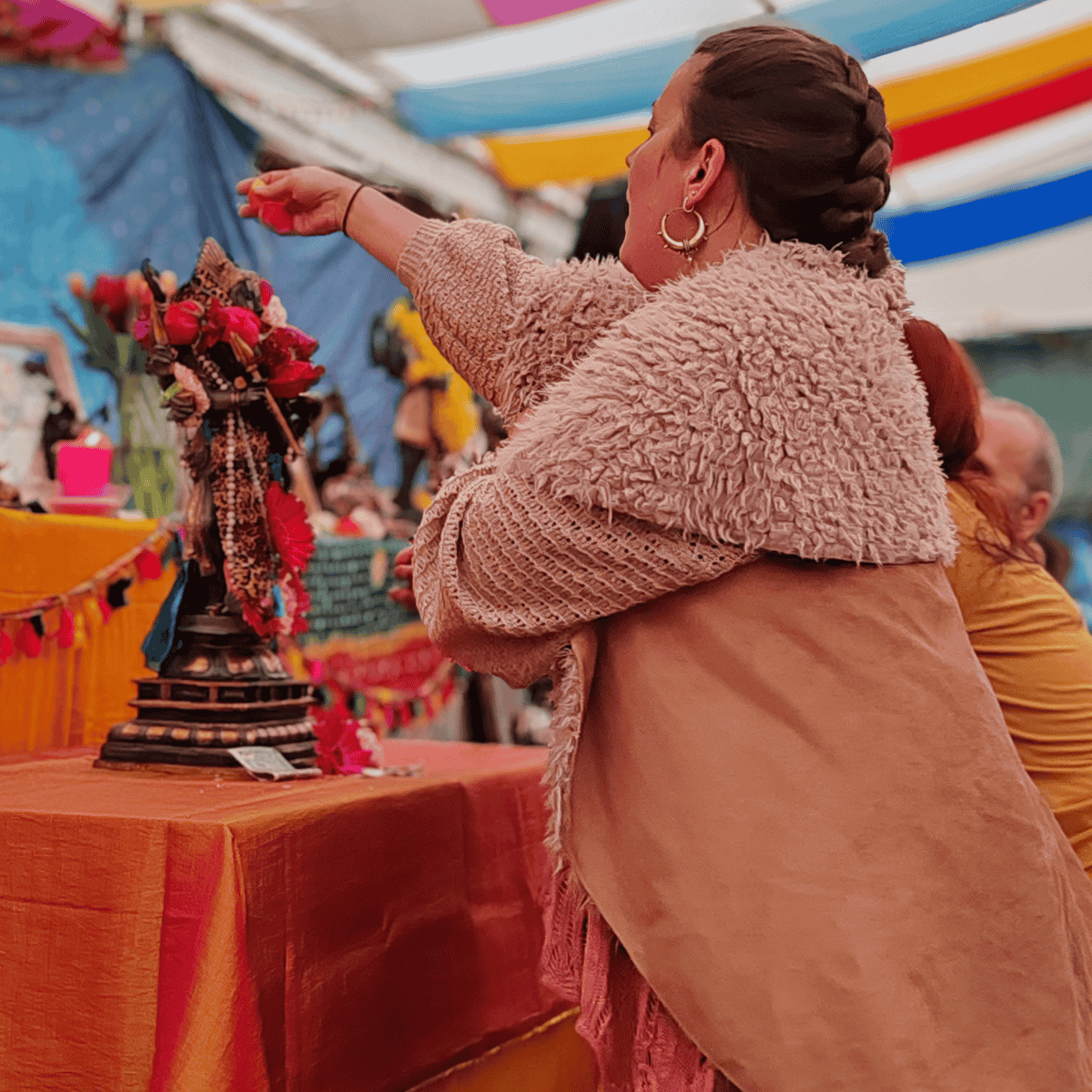 Woman with braided hair and hoop earrings wearing a cozy pink jacket at an outdoor market, reaching out towards colorful figurines and decorations on a table.