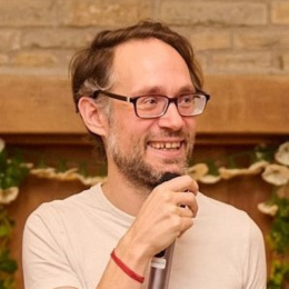 A man with glasses and a beard smiling, holding a microphone in a room with a brick wall and plants in the background.