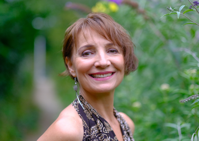 A portrait of a smiling woman with short, light brown hair, wearing earrings and a sleeveless patterned top, outdoors with a blurred green background of trees and plants.
