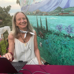 A smiling woman with long gray hair sitting at a table with a laptop, in front of a colorful outdoor mural of mountains and trees.