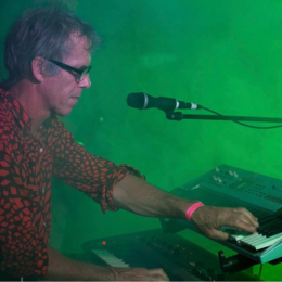 A man wearing glasses and a patterned shirt playing a keyboard on stage with green lighting.