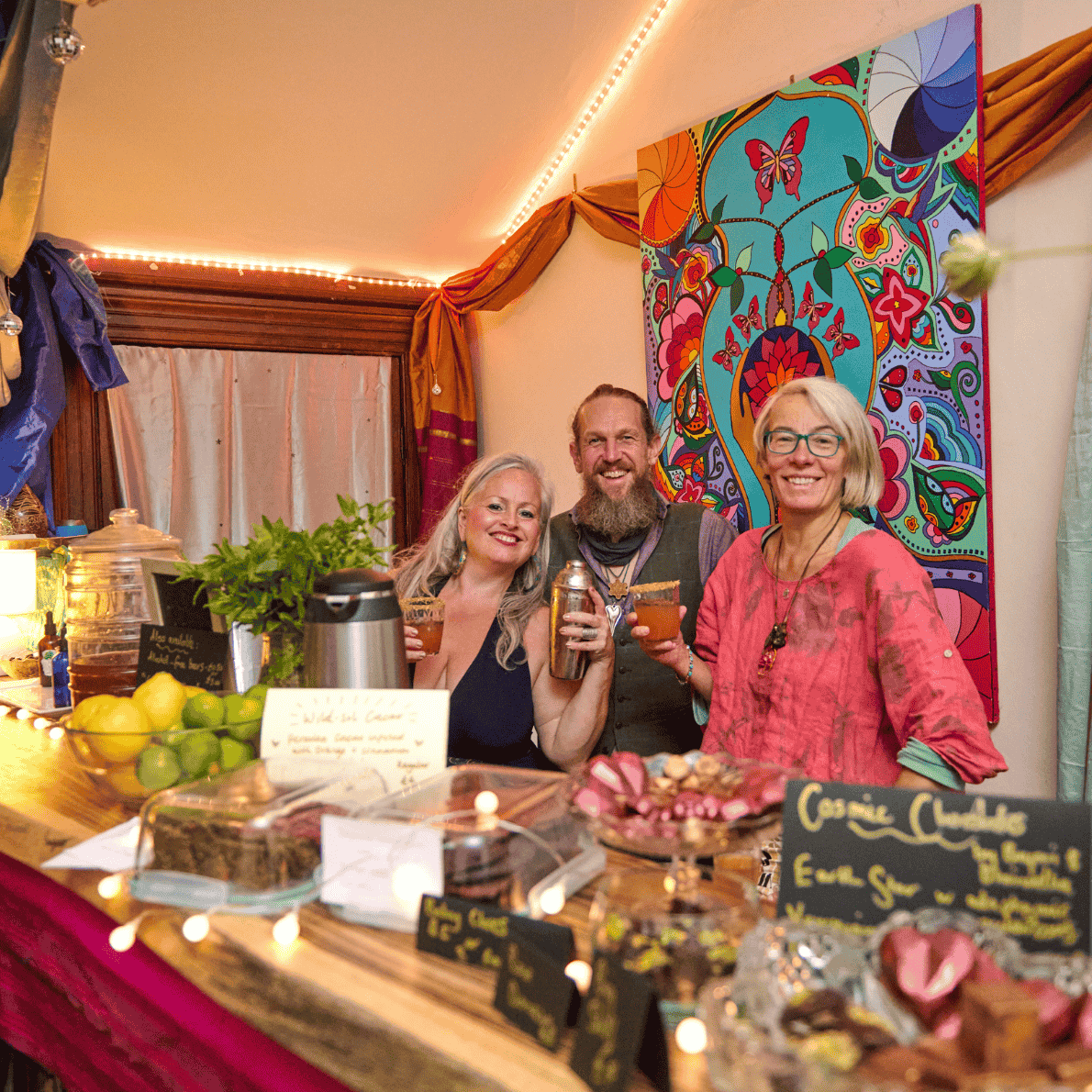 Three smiling people with drinks standing behind a colorful dessert and snack table at a party, with a vibrant, artistic background.