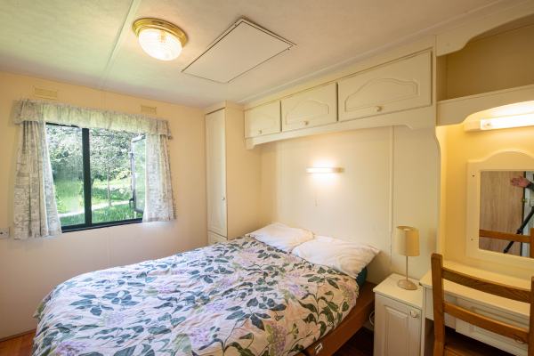 Bedroom with a bed, floral bedspread, two pillows, window with curtains, nightstand with a lamp, wall-mounted light, mirror with a wooden frame, and a chair with a wooden back.