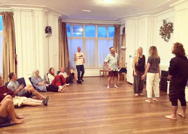 Group of adults participating in a workshop or class in a spacious room with wooden floors and large windows, some sitting on the floor and others standing and talking.
