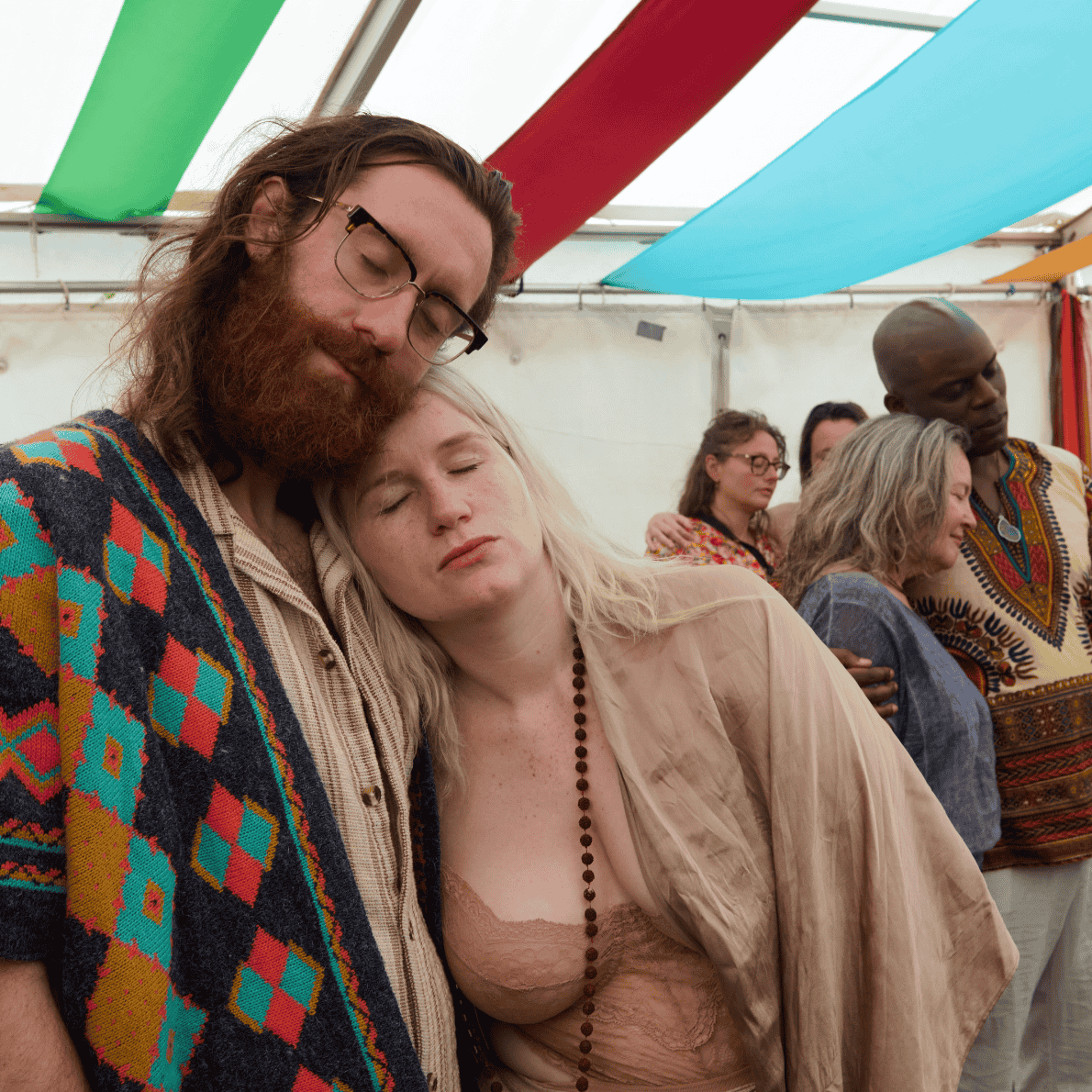 A group of people at a gathering under a colorful tent, with two individuals in the foreground resting their heads against each other, appearing peaceful or sleepy. The man has long hair, glasses, and a patterned sweater, while the woman has long blonde hair and is wearing a beige top and a beaded necklace.