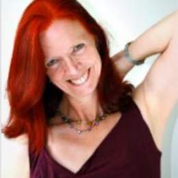 Woman with bright red hair smiling, wearing a dark purple top and jewelry