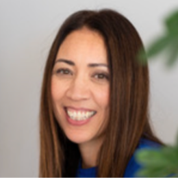 Smiling woman with long brown hair, wearing a blue top, against a light background with a blurred green plant in the foreground.