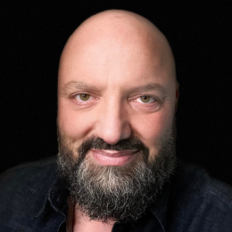 A close-up portrait of a bald man with a beard, smiling, against a black background.