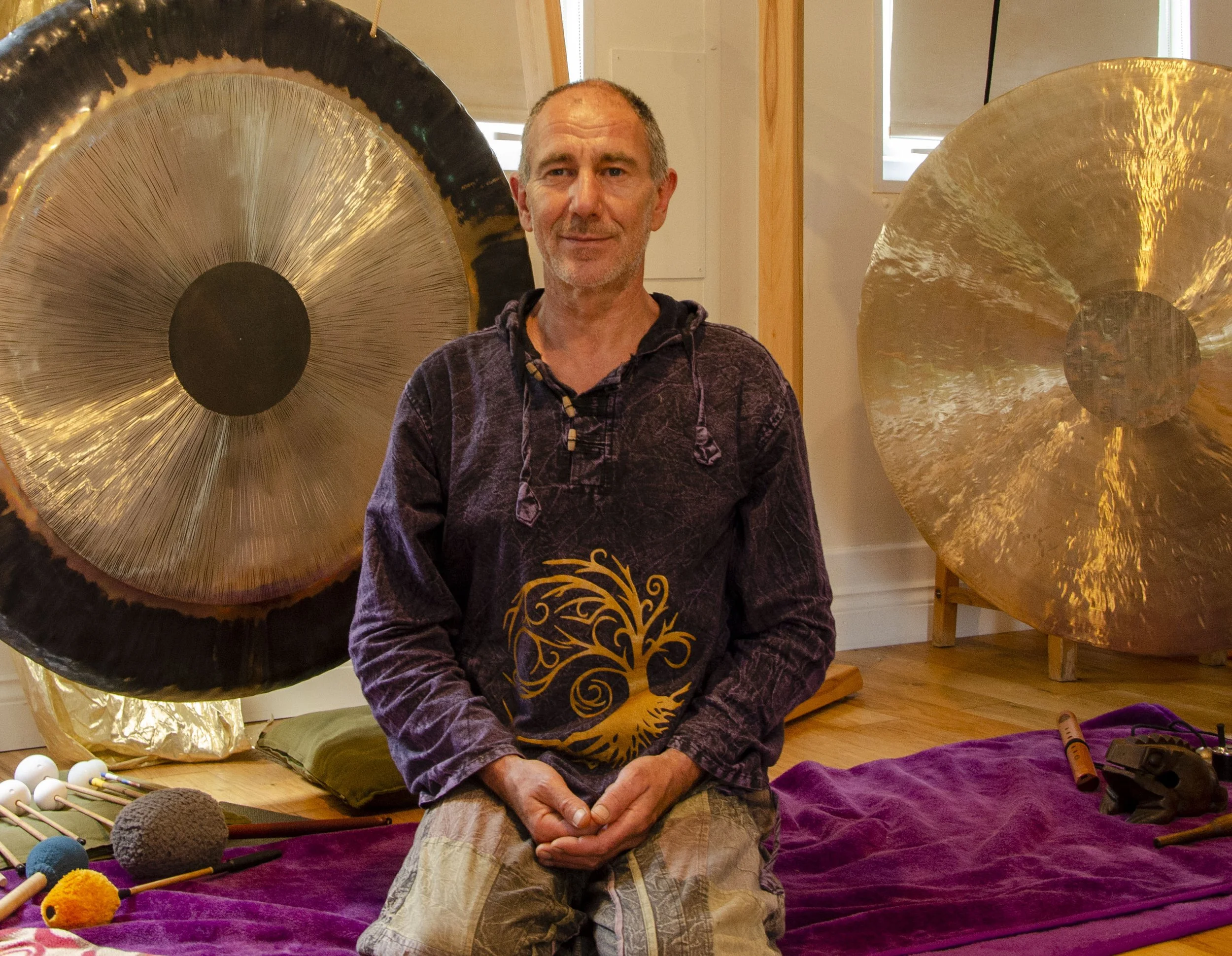 A man sitting cross-legged on a purple blanket, in front of large metallic gongs in a wooden-floored room.
