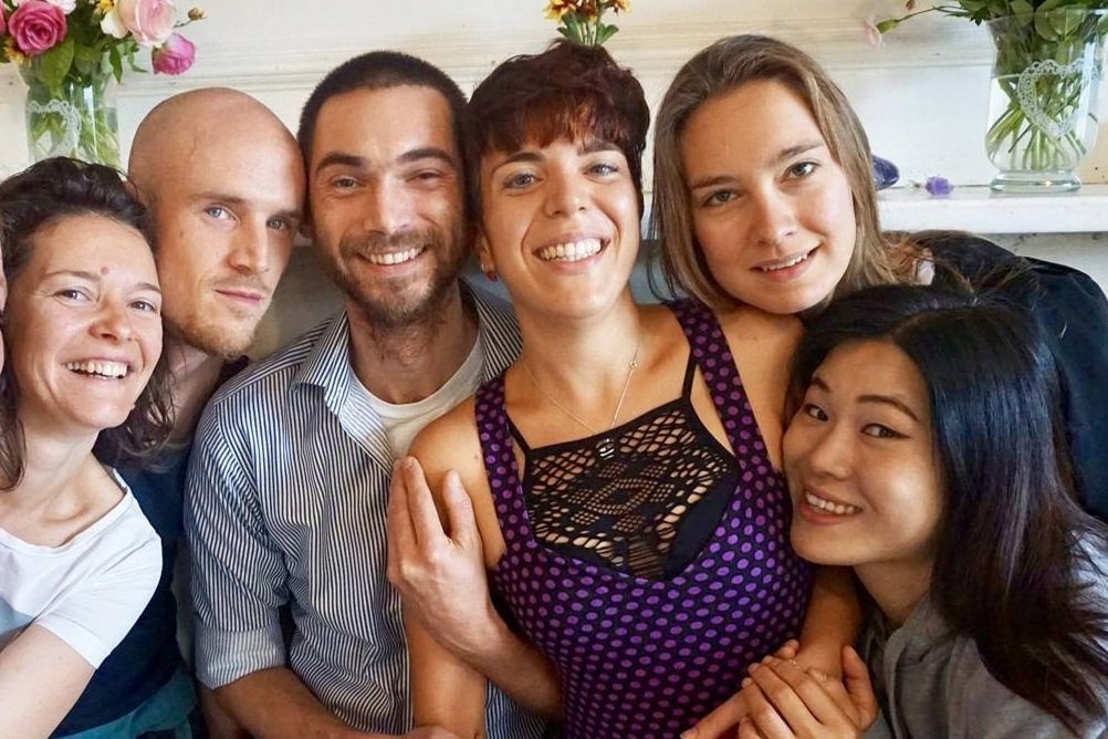 Six people smiling and posing closely together, with flowers in the background.