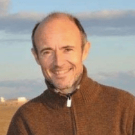 A smiling man with short hair and a beard, wearing a brown zip-up jacket, standing outdoors with a clear sky in the background.