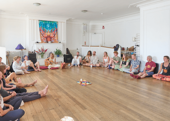 Group of people sitting in a circle on the floor of a spacious, white-walled room, with some playing guitar, participating in a group activity or meditation session.