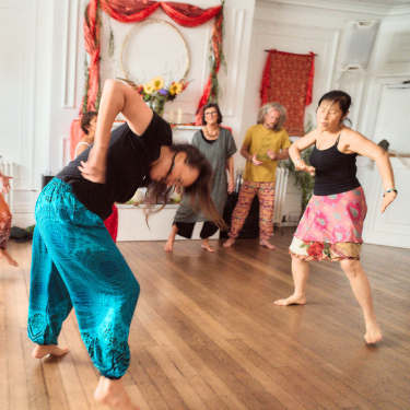 A group of women dancing in a brightly decorated room with white walls and wooden flooring, some wearing colorful skirts and tops.