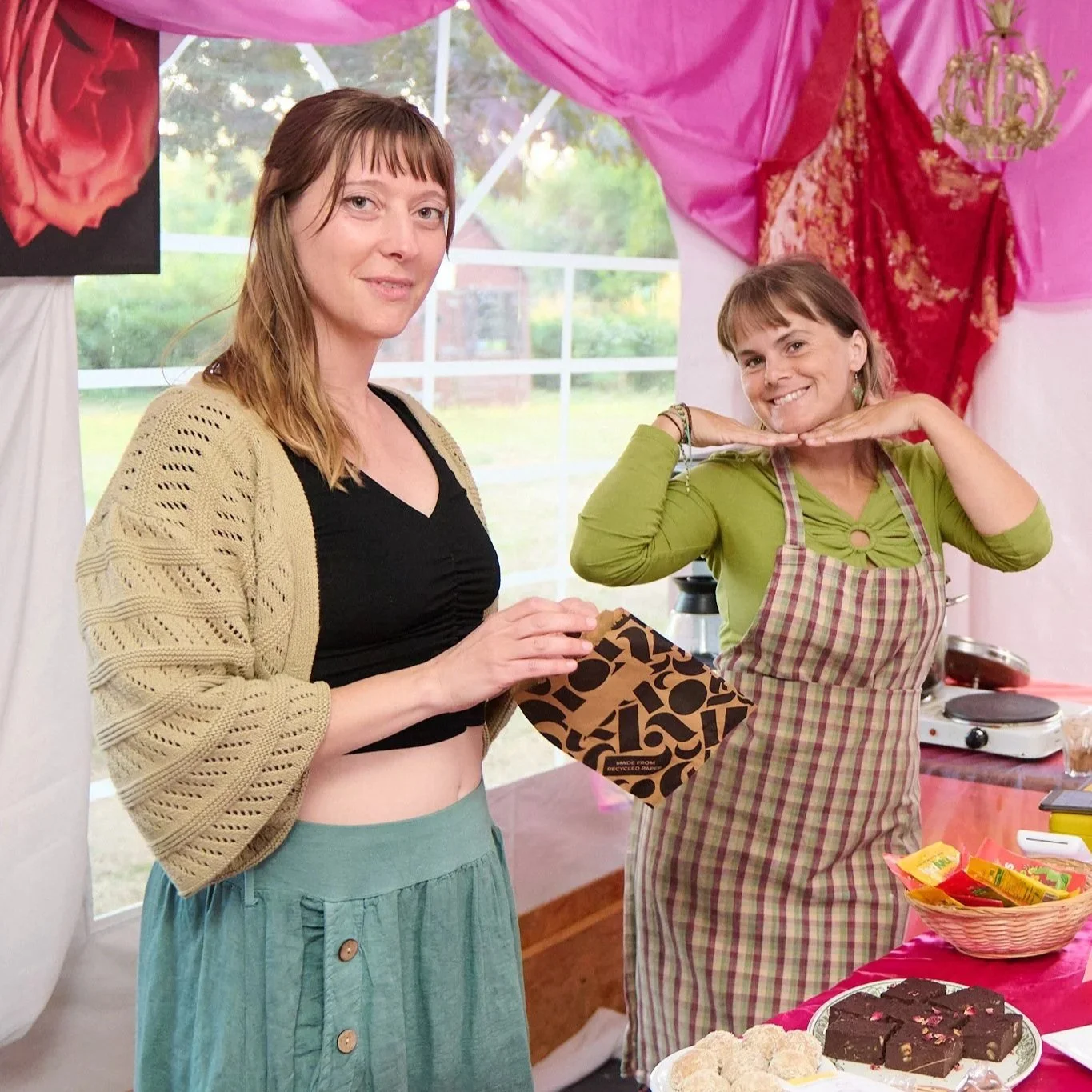 Two women at a party, one with brown hair wearing a beige cardigan and black top, holding a wrapped gift, and the other with brown hair in a green shirt and plaid apron, smiling and posing with hands under her chin. There is a table with snacks and a cake in front of them, decorated with pink and red drapes and a window with a green outdoor view.
