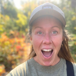 A woman outdoors with a surprised or excited expression, wearing a cap and casual clothing, with trees and autumn foliage in the background.