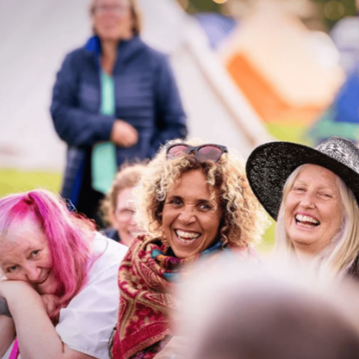 Group of smiling women enjoying a sunny outdoor event, with colorful tents visible in the background.