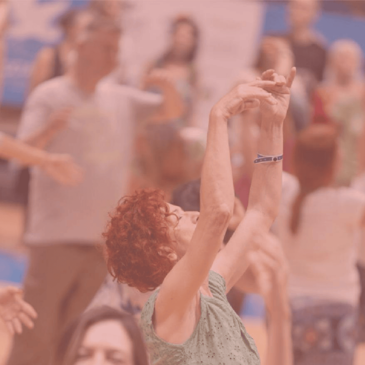 A woman with curly red hair is dancing with her arms raised in a gymnasium or dance studio, surrounded by other people also dancing or exercising.