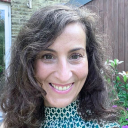 Smiling woman with wavy dark hair outdoors, wearing a patterned top, in front of a wooden fence and some greenery.