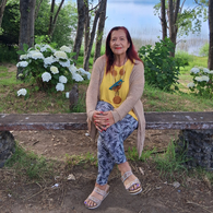 Woman sitting on a stone bench outdoors with trees and hydrangea flowers in the background, wearing a yellow top and patterned pants.