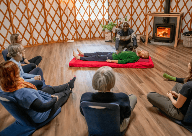 An indoor workshop or class with an instructor demonstrating a hands-on activity to a group of seated women, with one woman lying on a red mat and the instructor performing a technique on her, in a cozy room with wooden floors, a fireplace, and decorative lattice walls.