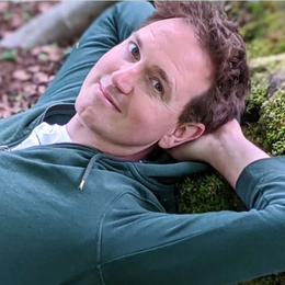 A man lying on the grass outdoors, resting his head on his hand and looking at the camera.
