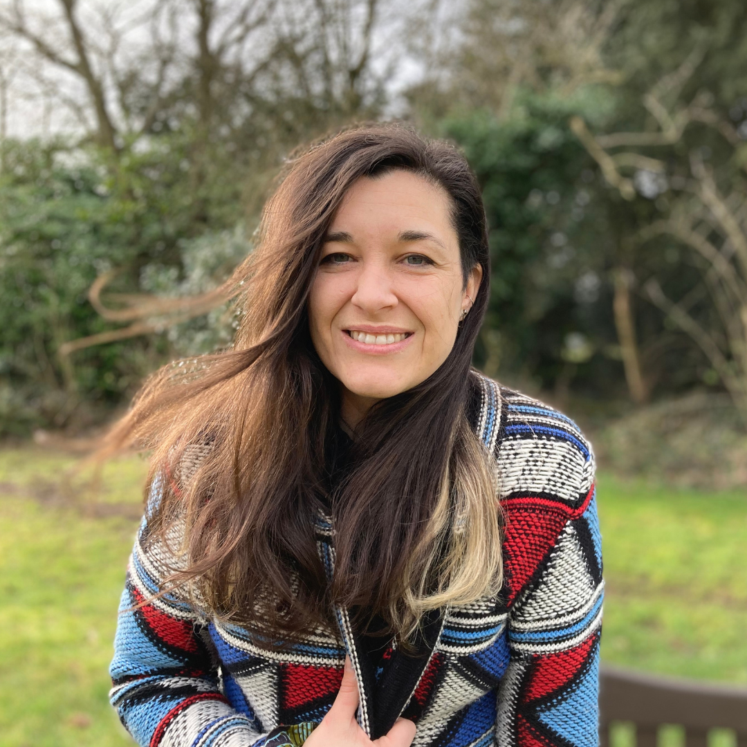 Woman with long dark hair wearing a colorful patterned jacket smiling outdoors in a garden with trees in the background.