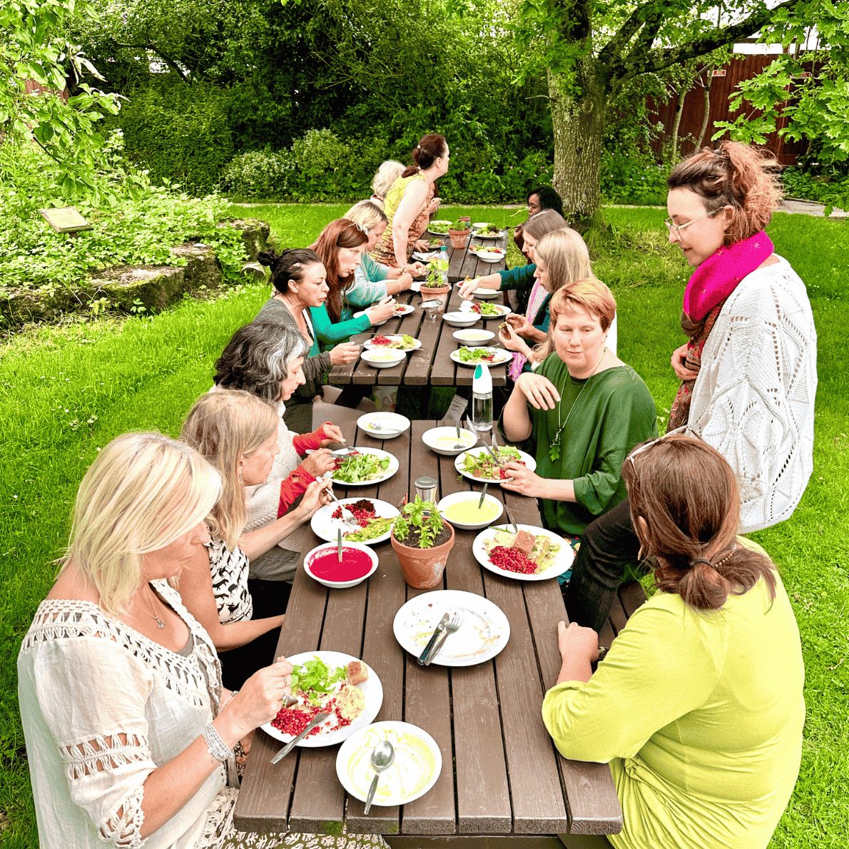 A group of women having a meal outdoors at a long wooden table in a lush green garden, with salads and bowls of soup.