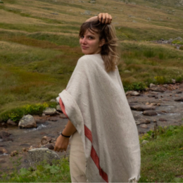 Young woman with brown hair in an outdoor grassy field, wearing a beige poncho and holding her right arm over her head, looking back at the camera.