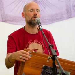 A man with glasses and a beard, wearing a red T-shirt, plays a kalimba while speaking into a microphone against a white background with a purple decorative cloth.